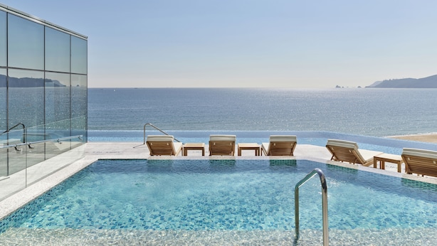 A pool with clear blue water, wooden lounge chairs, and a glass wall overlooking the ocean. - Grand Josun Busan (Busan)