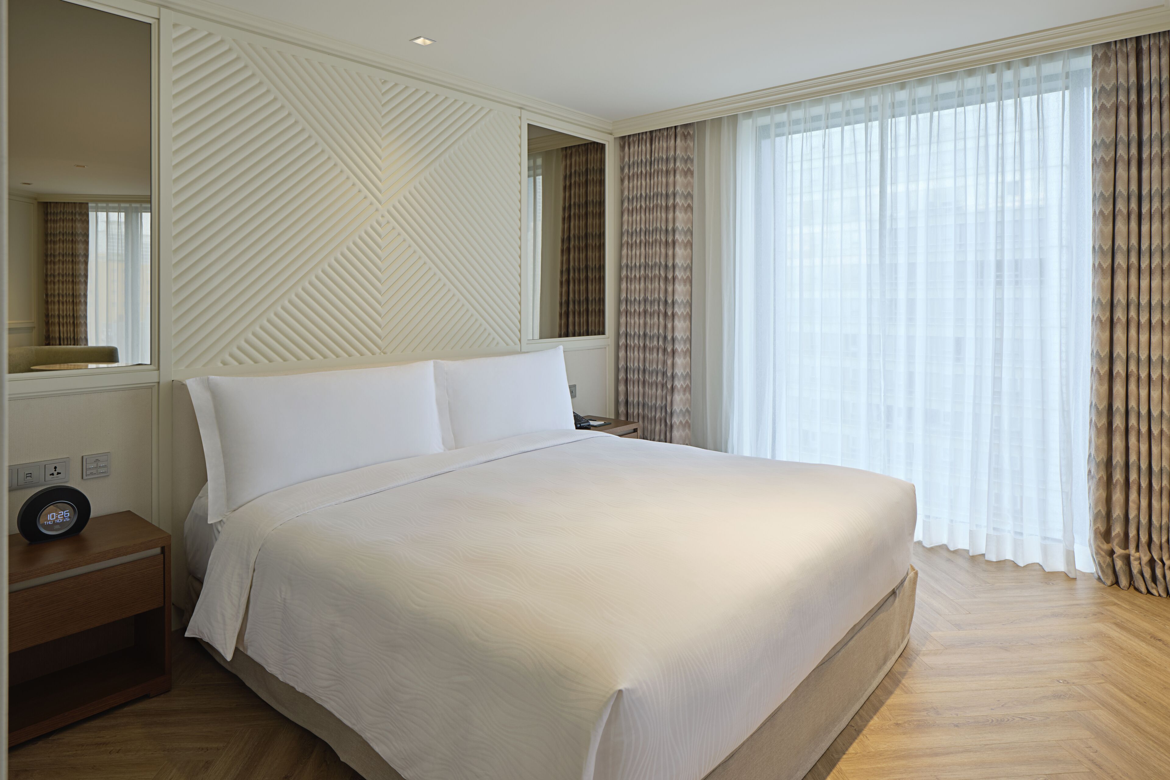 A neatly made bed in a hotel room with a large mirror, a bedside table with a clock, and a window with curtains.