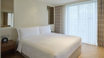 A neatly made bed in a hotel room with a large mirror, a bedside table with a clock, and a window with curtains.