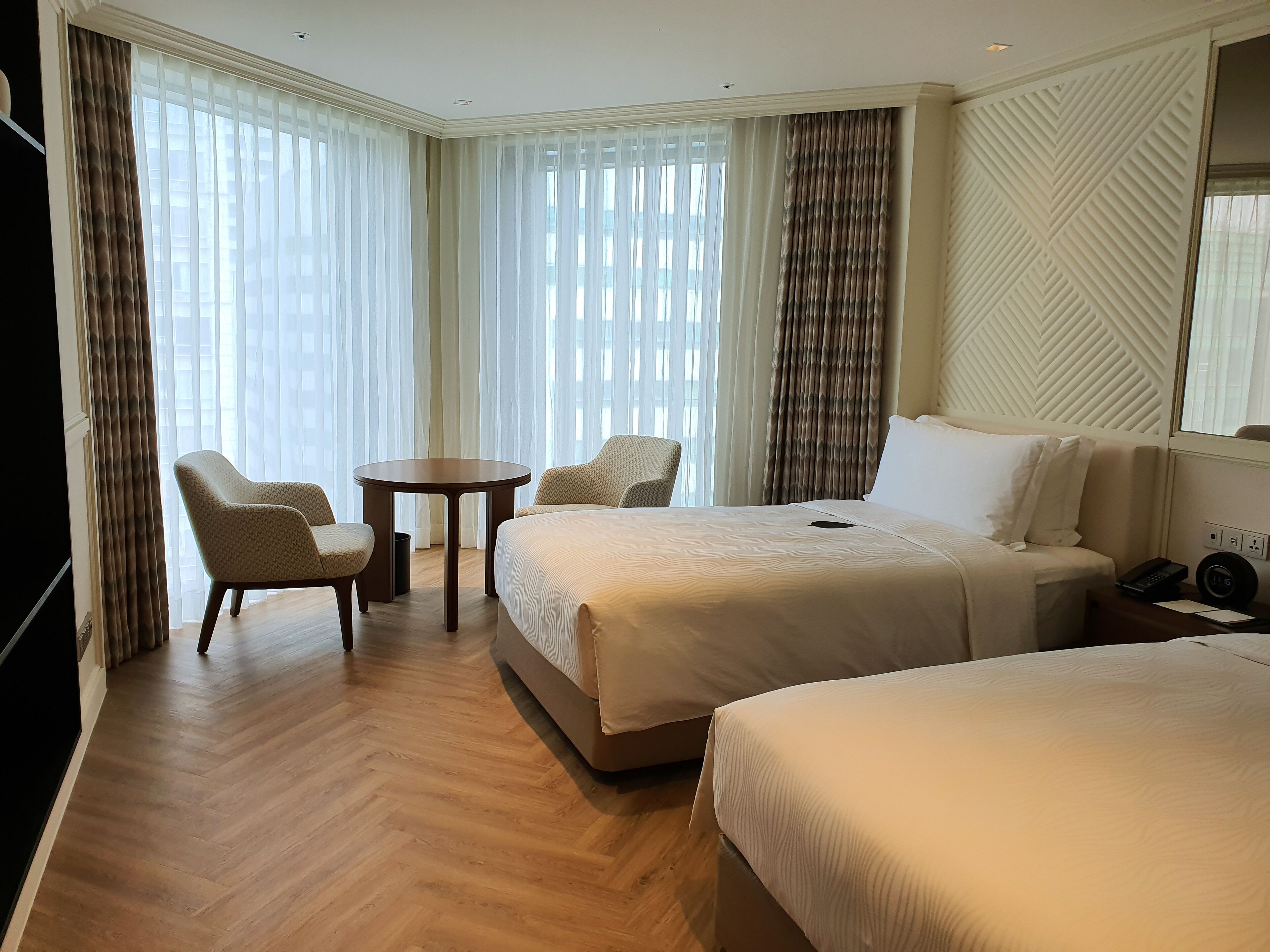 A hotel room with two beds, a small table, two chairs, a TV, and a large window with curtains.