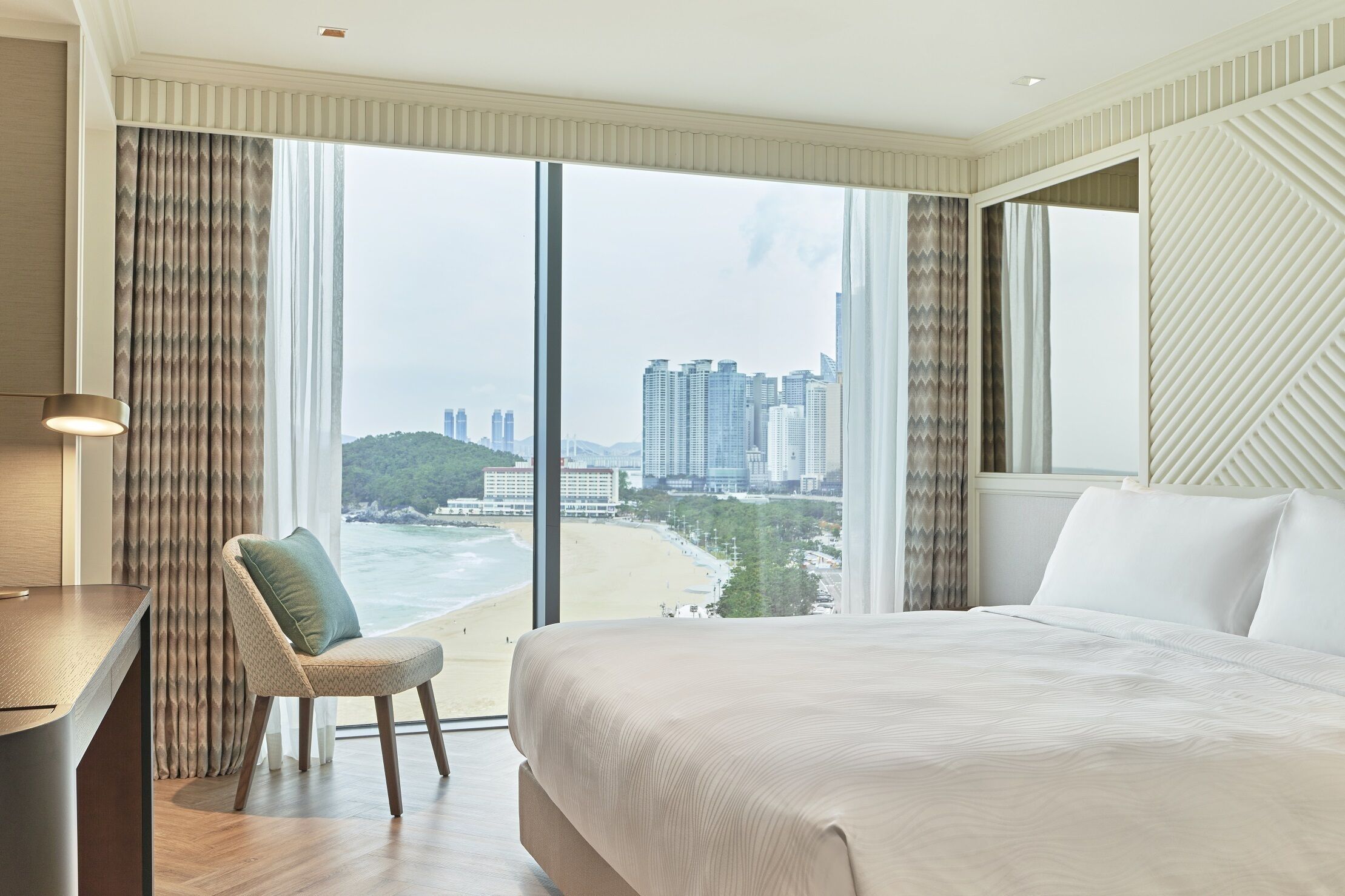 Habitación de hotel con una cama grande, un escritorio y una silla. La habitación tiene vistas a la playa y al paisaje urbano.