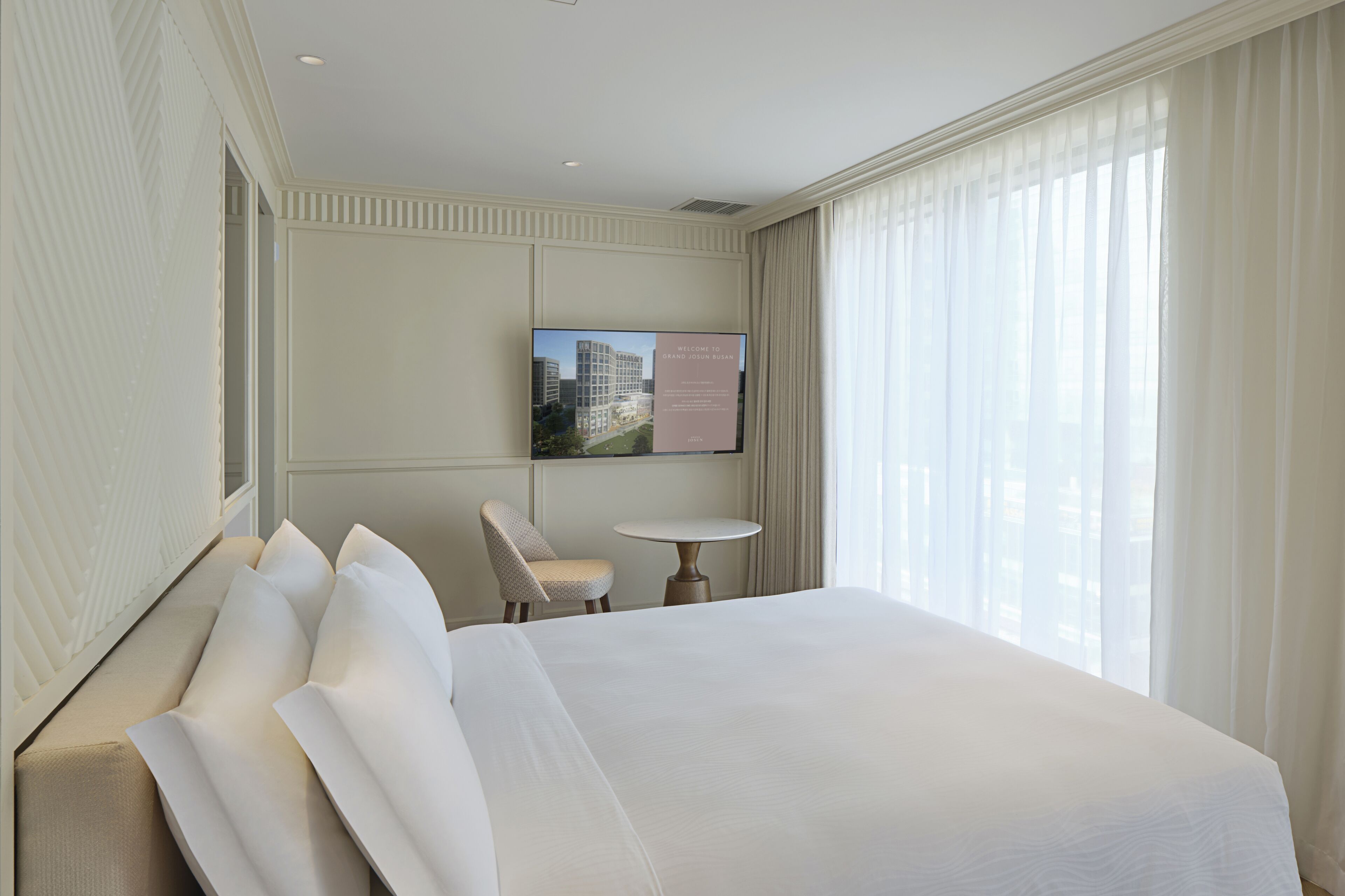 Quarto de hotel com uma cama grande, uma mesinha, uma cadeira e uma televisão na parede exibindo uma mensagem de boas-vindas.