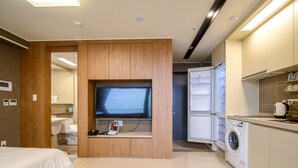 A modern hotel room with a flat-screen TV mounted on a wooden panel, a bed, a washing machine, and a kitchenette. - Sokcho Blue Terra Pension (Sokcho)