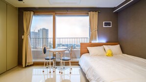 A hotel room with a bed, a small table with chairs, a window with curtains, and a city view. - Sokcho Blue Terra Pension (Sokcho)
