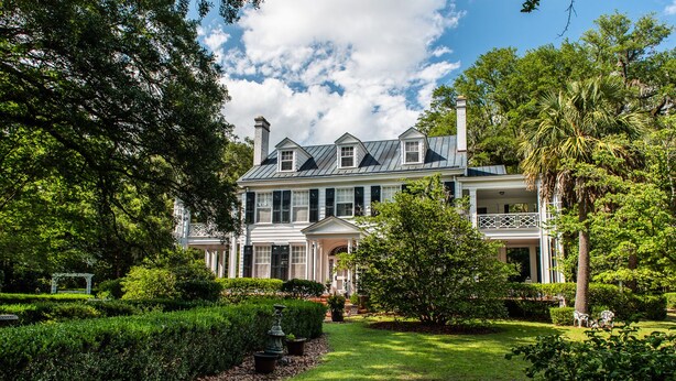 A large, two-story house with a symmetrical facade, a central entrance with a portico, and a well-manicured garden with a lawn, hedges, and a few trees. - Winter Discounts/ We love pets and they love our amenities (Summerville)
