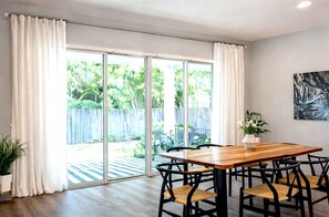 Un coin repas avec une table en bois et des chaises noires, de grandes portes-fenêtres coulissantes donnant sur un jardin, et une plante en pot sur la gauche.