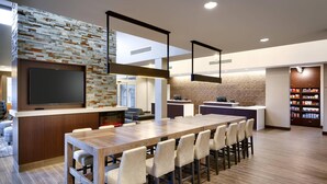 A modern kitchen with a long wooden table, bar stools, a built-in TV, and a brick accent wall.