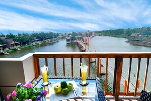 Un balcon avec une table, deux boissons et une assiette de fruits. On aperçoit, au loin, une rivière avec des bateaux et un pont.