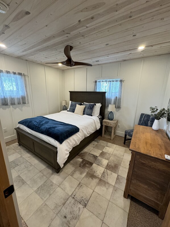 A bedroom with a bed, bedside tables, a wooden dresser, a ceiling fan, and a window with curtains.