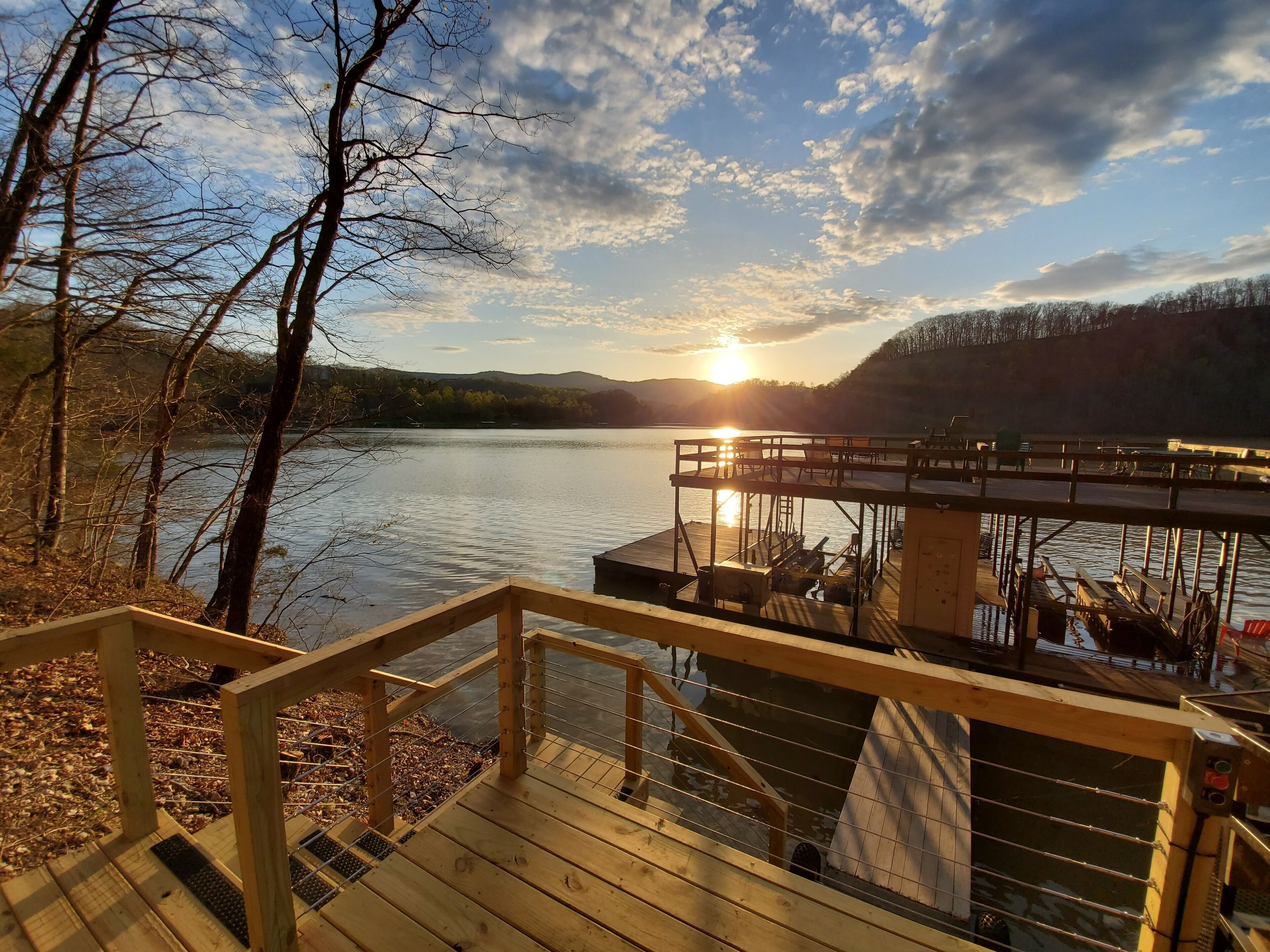 A wooden deck with a railing overlooking a calm lake, a dock with boats, and a sunset in the background.