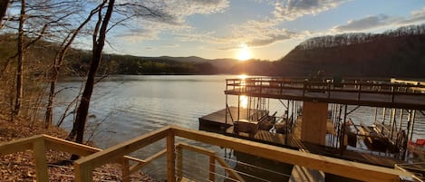 A wooden deck with a railing overlooking a calm lake, a dock with boats, and a sunset in the background.