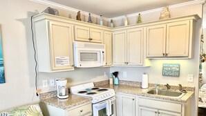 A compact kitchen with white cabinets, a microwave, stove, and a sink. There is a coffee maker, a trash bin, and a decorative sign on the wall. - 2nd Floor Beachside Condo•Private Beach Access•Pool•Kitchen•Pet-Friendly (Atlantic Beach)