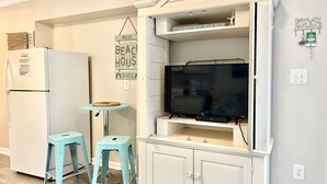 A compact kitchen with a white refrigerator, a small teal table, two matching stools, a TV on a white cabinet, and a 'Keys' sign on the wall. - 2nd Floor Beachside Condo•Private Beach Access•Pool•Kitchen•Pet-Friendly (Atlantic Beach)