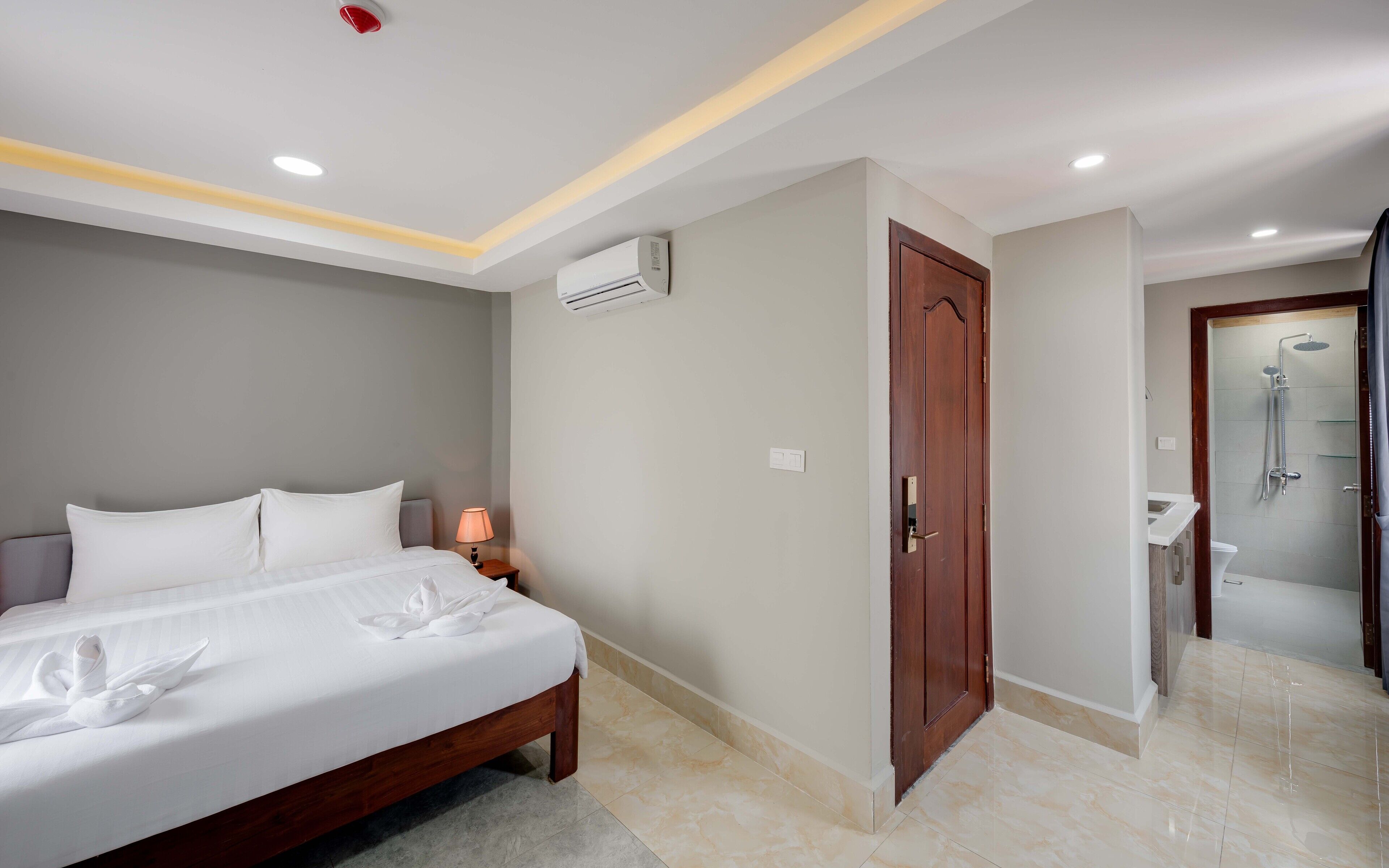 A hotel room with a bed, a nightstand, an air conditioning unit, a wooden door, and a shower area.