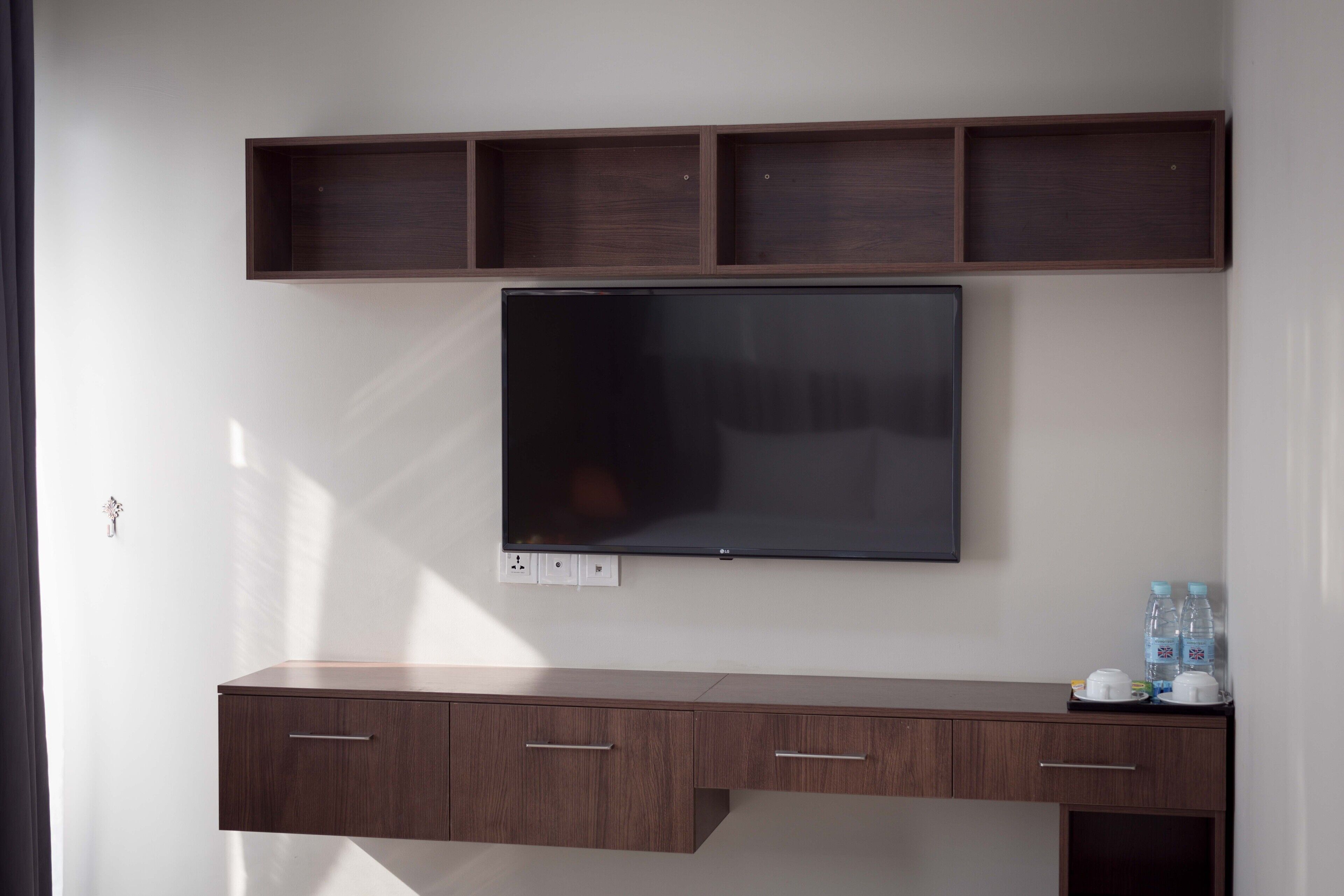 A modern room with a flat-screen TV mounted on the wall, a wooden desk with drawers, and a shelf above the TV.