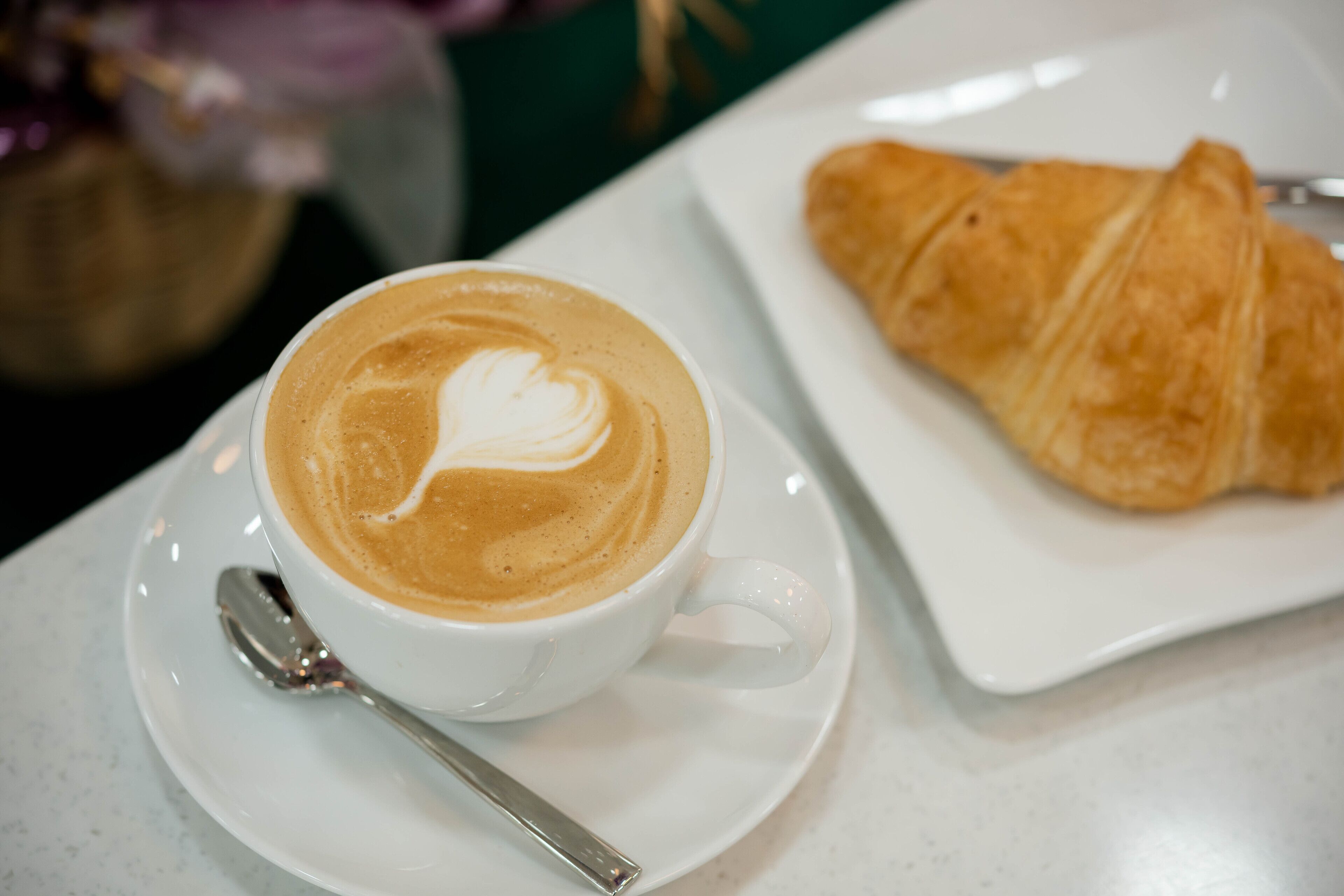 A cup of coffee with latte art on a white saucer, accompanied by a croissant on a white plate.