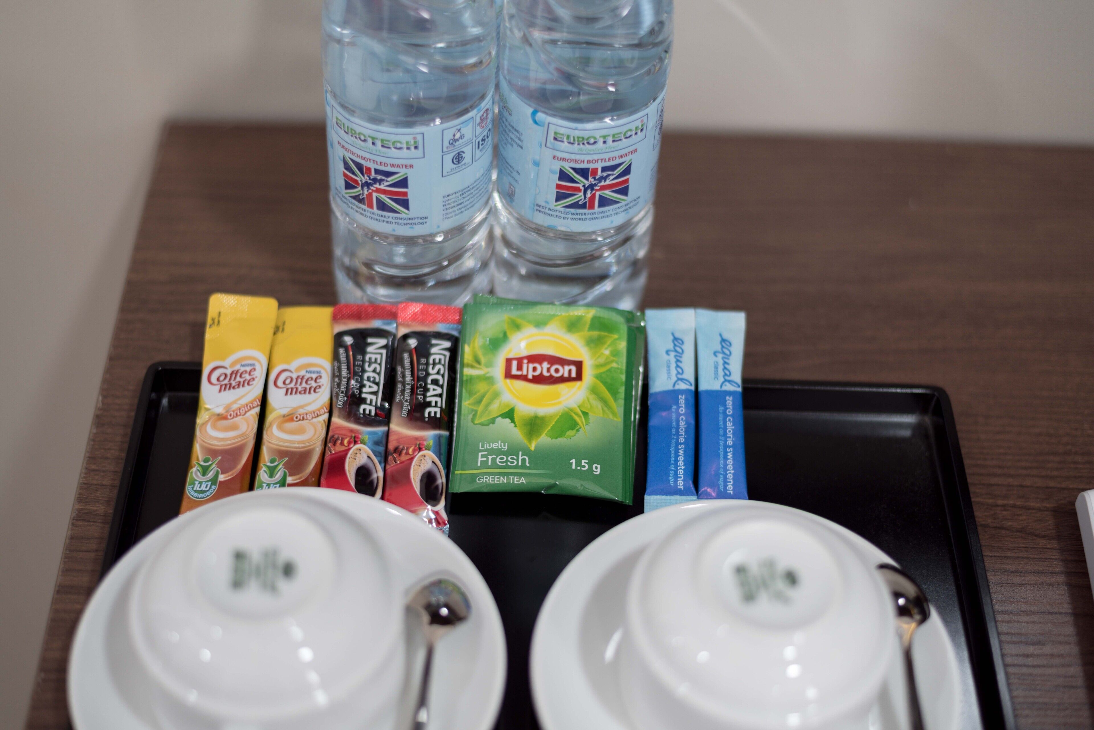 Una bandeja con dos tazas blancas, té verde Lipton, sobres de café y agua embotellada.
