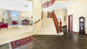 A spacious living area with a grand staircase, a piano, and a large window offering a view. - Razorback Dream Farmhouse: 10BR, Space & Serenity (Razorback)