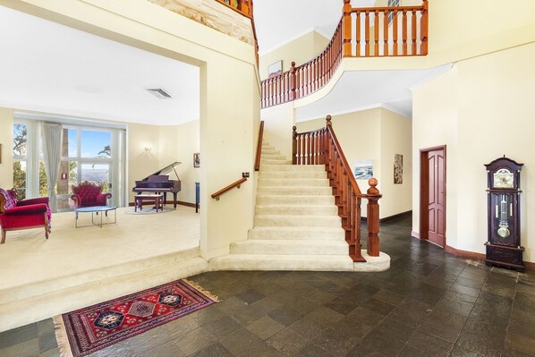 A spacious living area with a grand staircase, a piano, and a large window offering a view. - Razorback Dream Farmhouse: 10BR, Space & Serenity (Razorback)