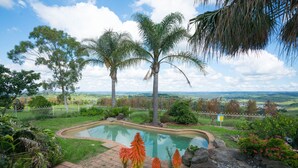 A landscaped garden with a swimming pool, palm trees, and a view of distant hills. - Razorback Dream Farmhouse: 10BR, Space & Serenity (Razorback)
