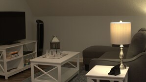 A living room with a TV, a lamp, a small table with a decorative item, a bookshelf, and a patterned rug.