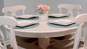 A round dining table with white chairs, a white vase with flowers, and blue placemats.