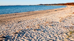 Ein sandiger Strand mit klarem, blauem Wasser und sanften Hügeln im Hintergrund.
