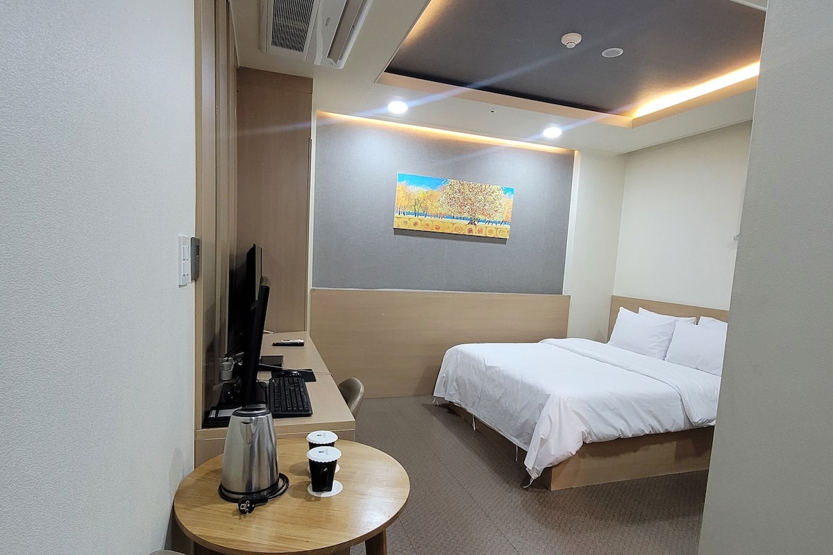 A hotel room with a bed, a desk with a coffee maker, a chair, and a painting on the wall.