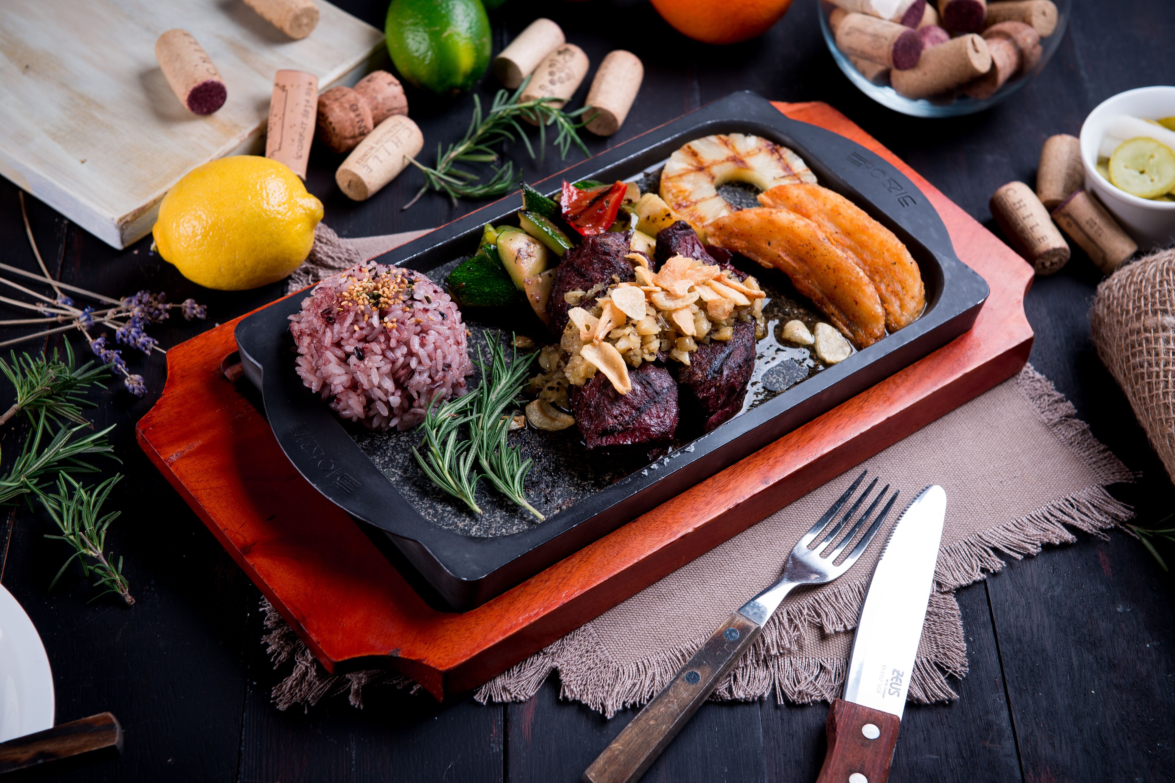 A black slate serving tray with a variety of food items including rice, meat, and vegetables, accompanied by a knife and fork on a wooden board.