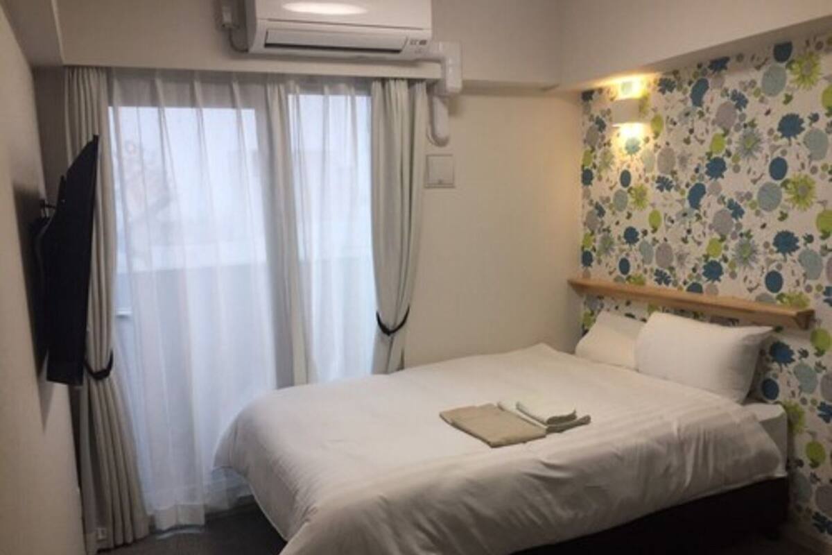 A hotel room with a bed, air conditioning unit, curtains, and a floral wallpaper.