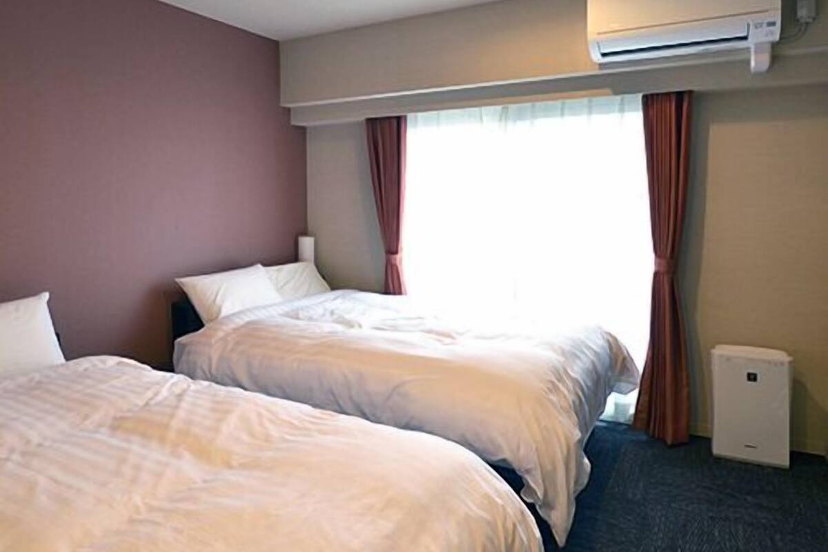 A hotel room with two beds, an air conditioning unit, a window with curtains, and a small bedside table.
