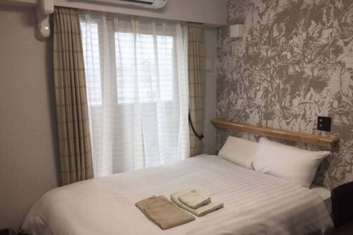 A hotel room with a bed, a wooden headboard, a window with sheer curtains, and a wall with a floral pattern.