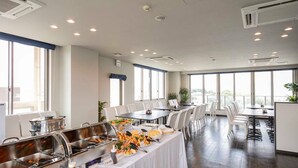 A buffet setup with various food items, a white tablecloth-covered table, and a modern interior with large windows and ceiling lights. - Omura Station Hotel Nagasaki Airport (Omura)