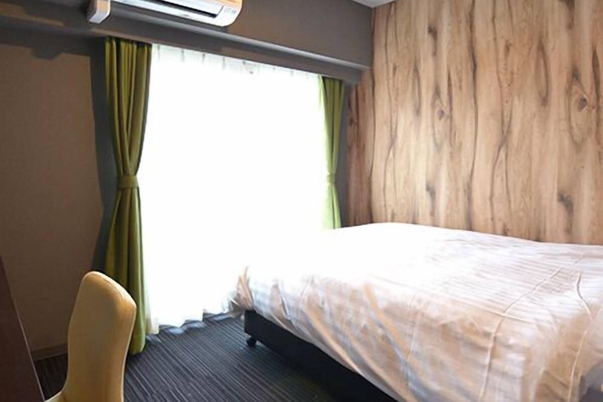 A hotel room with a bed, a chair, an air conditioning unit, and a window with curtains.