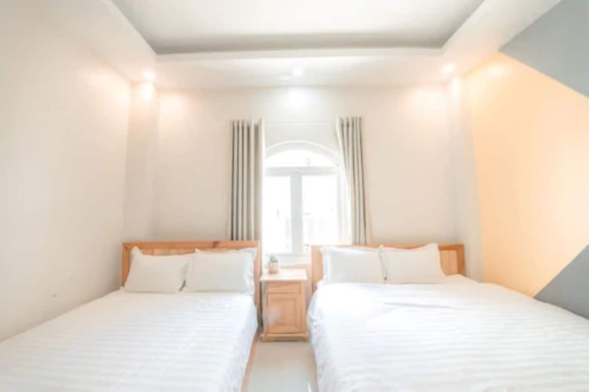 A hotel room with two single beds, a small bedside table, and a view of the street through a window with curtains.