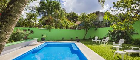 A rectangular swimming pool surrounded by a green wall, with white lounge chairs and a grassy area with trees and plants.