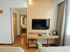 A hotel room with a bed, a desk with a television, a trash bin, and a window with curtains.