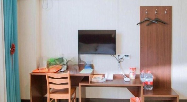 A hotel room with a wooden desk, a television mounted on the wall, a chair, and a wardrobe with hangers.