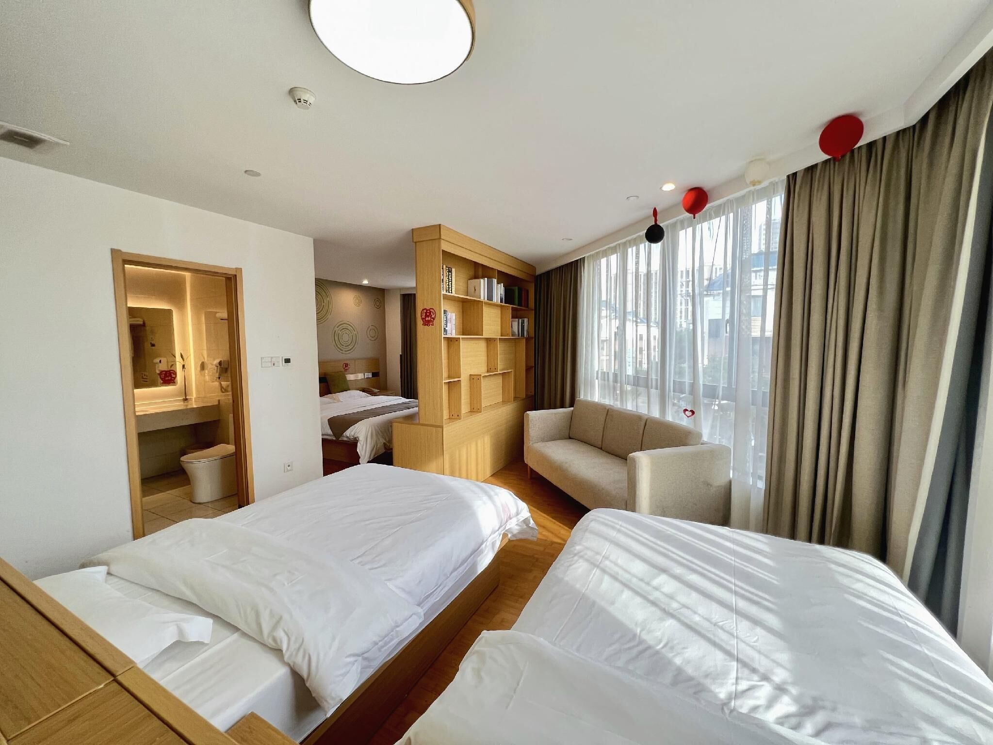 A hotel room with two beds, a wooden wardrobe, a small sofa, and a large window with curtains.