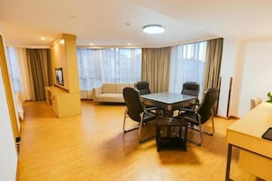 Habitación de hotel con piso de madera, una mesa redonda con cuatro sillas, un sofá y un amplio ventanal con cortinas.