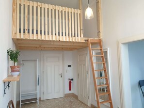 A room with a lofted sleeping area, a wooden ladder, a hanging light fixture, a door, and a small shelf with a plant.
