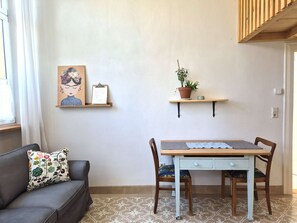 A room with a grey sofa, a wooden table with two chairs, a wall-mounted shelf with a plant, and a framed artwork.