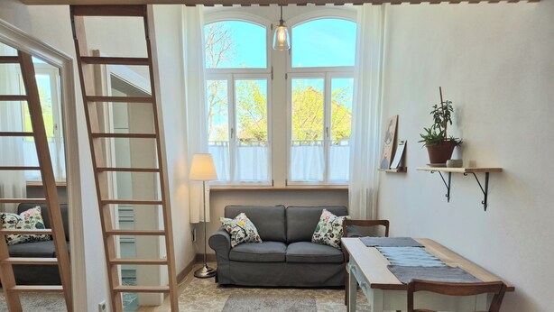 Ein Zimmer mit einer Couch, einem Tisch, einer Hockerleiter, einer Stehlampe und einem Fenster mit Blick auf Bäume.