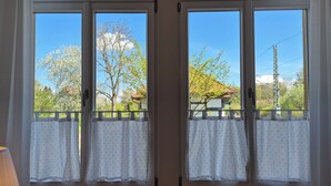 Ein Zimmer mit zwei Fenstern, jeweils mit weißem Vorhang und Blick auf Bäume und ein Gebäude.