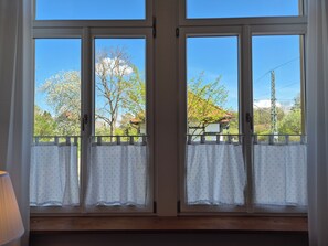 Habitación con dos ventanas, cada una con cortina blanca y vistas a árboles y un edificio.