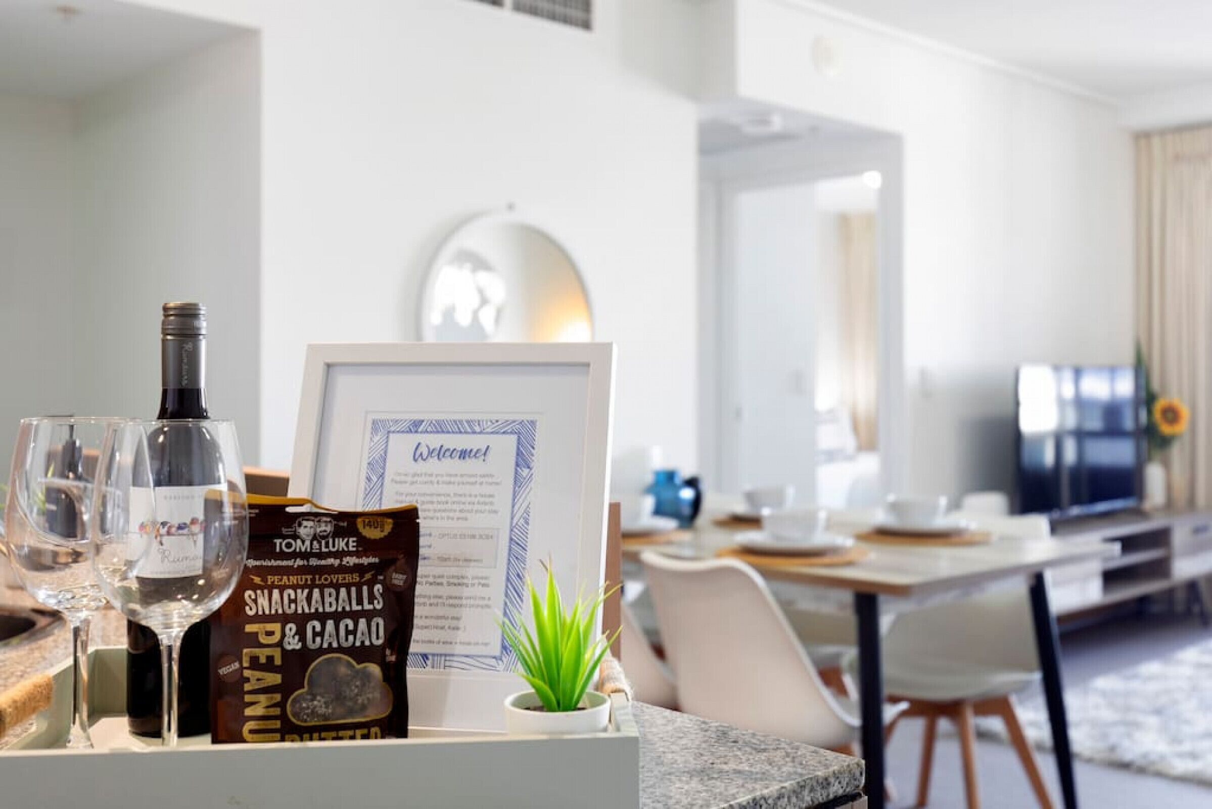 A hotel room with a dining area, a framed welcome sign, a bottle of wine, a snack bar sign, and a small plant on the counter.