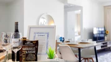 A hotel room with a dining area, a framed welcome sign, a bottle of wine, a snack bar sign, and a small plant on the counter.