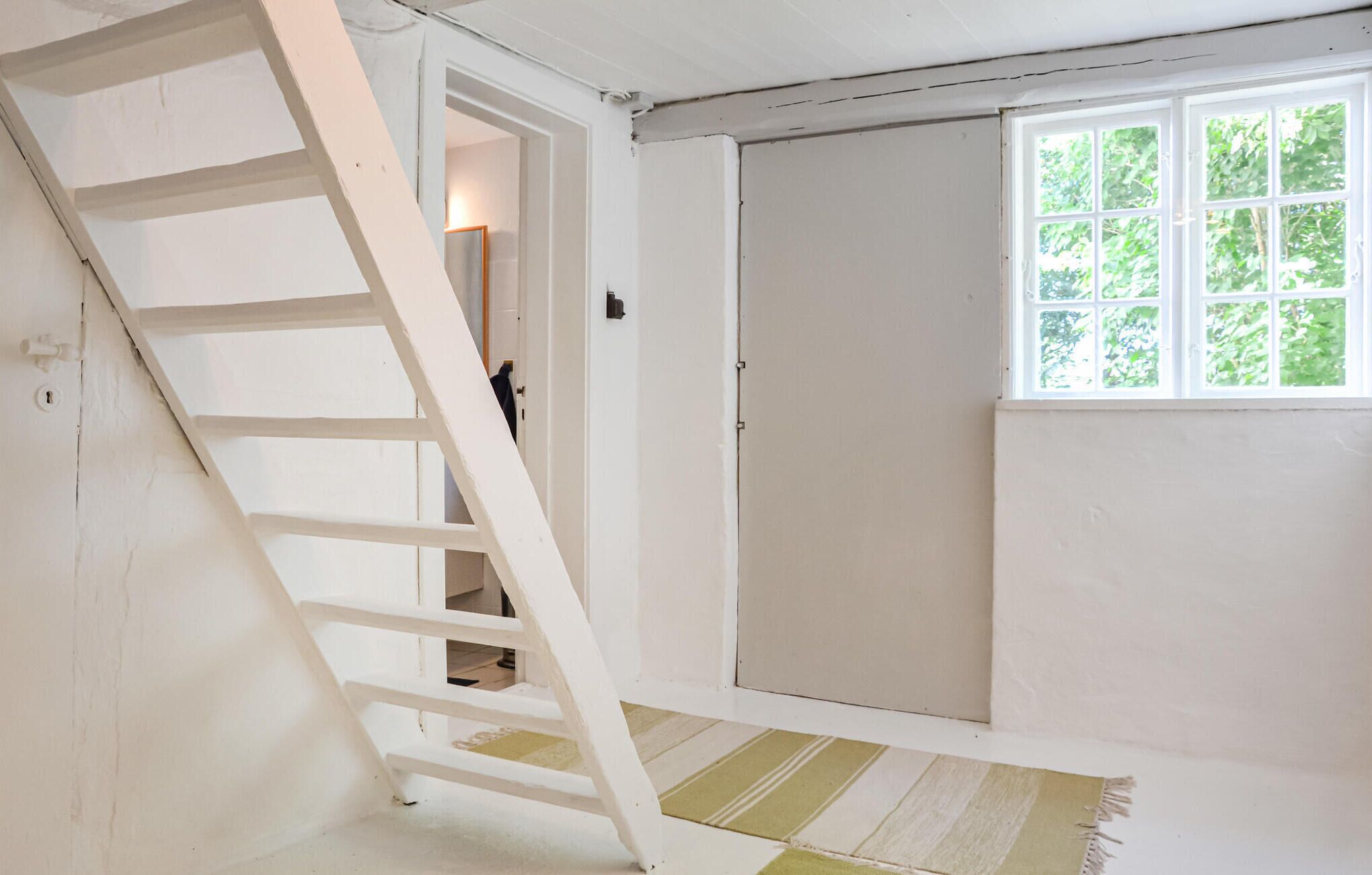A room with a white door, a window with a view of greenery, and a white ladder leaning against the wall.