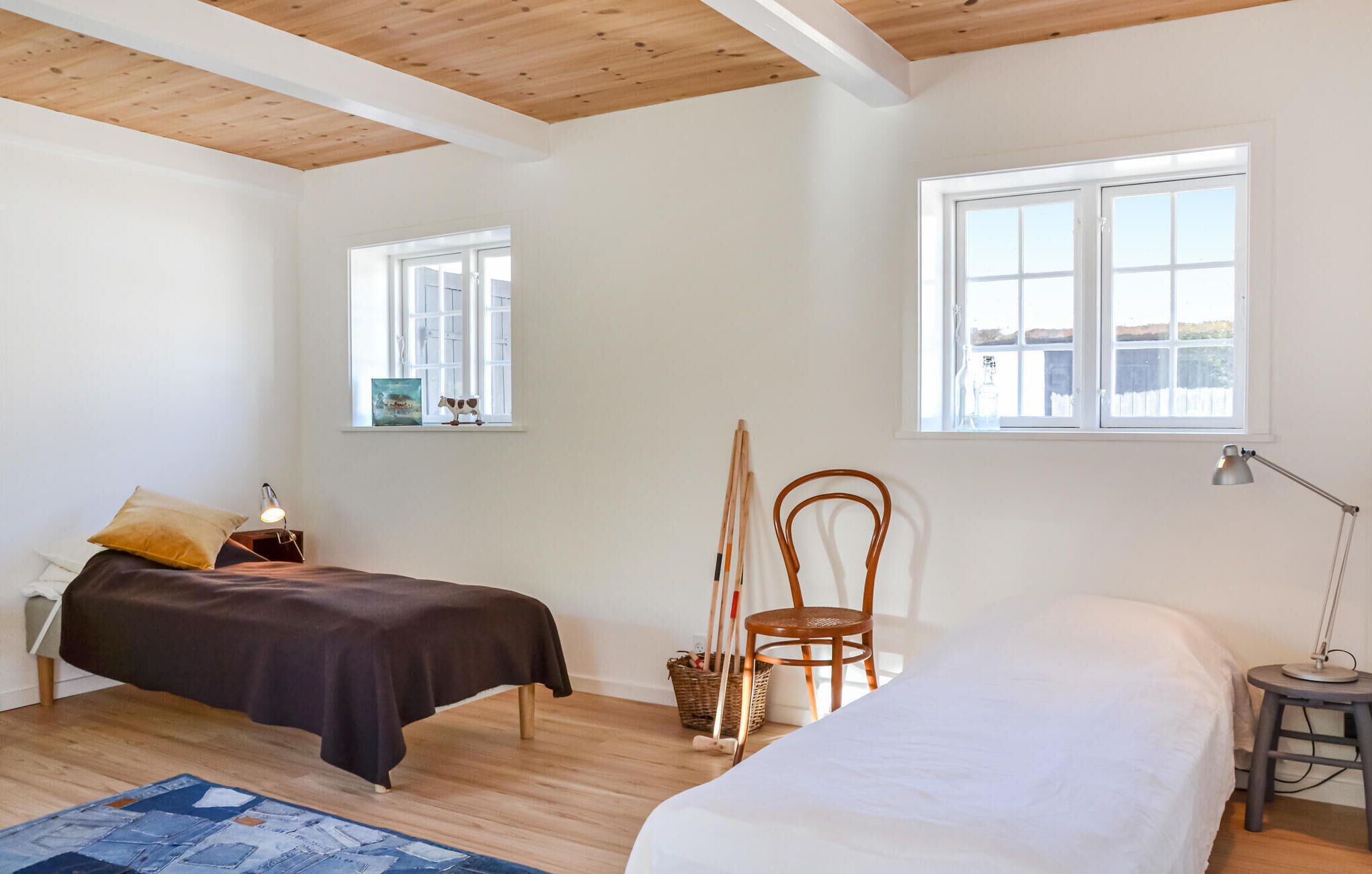 Une chambre avec deux lits, un plancher en bois, un plafond avec des poutres apparentes et deux fenêtres laissant entrer la lumière naturelle.