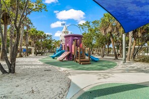 Explore a vibrant play area with a multi-level play structure, slides, and climbing elements, surrounded by palm trees and a sandy ground.